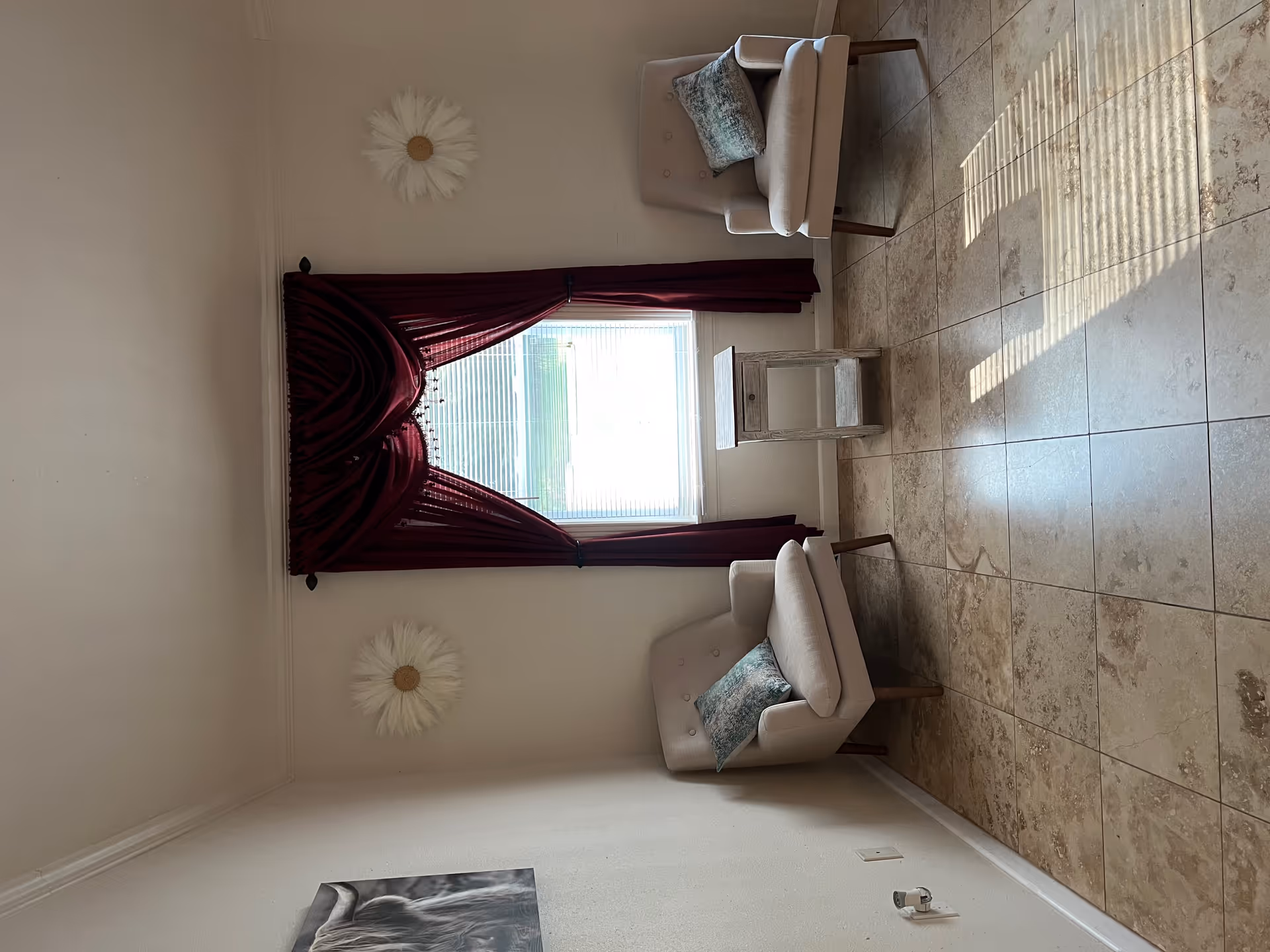 A small sitting area with two beige armchairs, each with a decorative pillow, positioned on either side of a small wooden side table. Behind the table is a window with closed blinds and burgundy curtains. The walls are light-colored and decorated with two white flower wall hangings and a black and white picture of a cow on the left wall. The floor is tiled with a beige and brown pattern.
