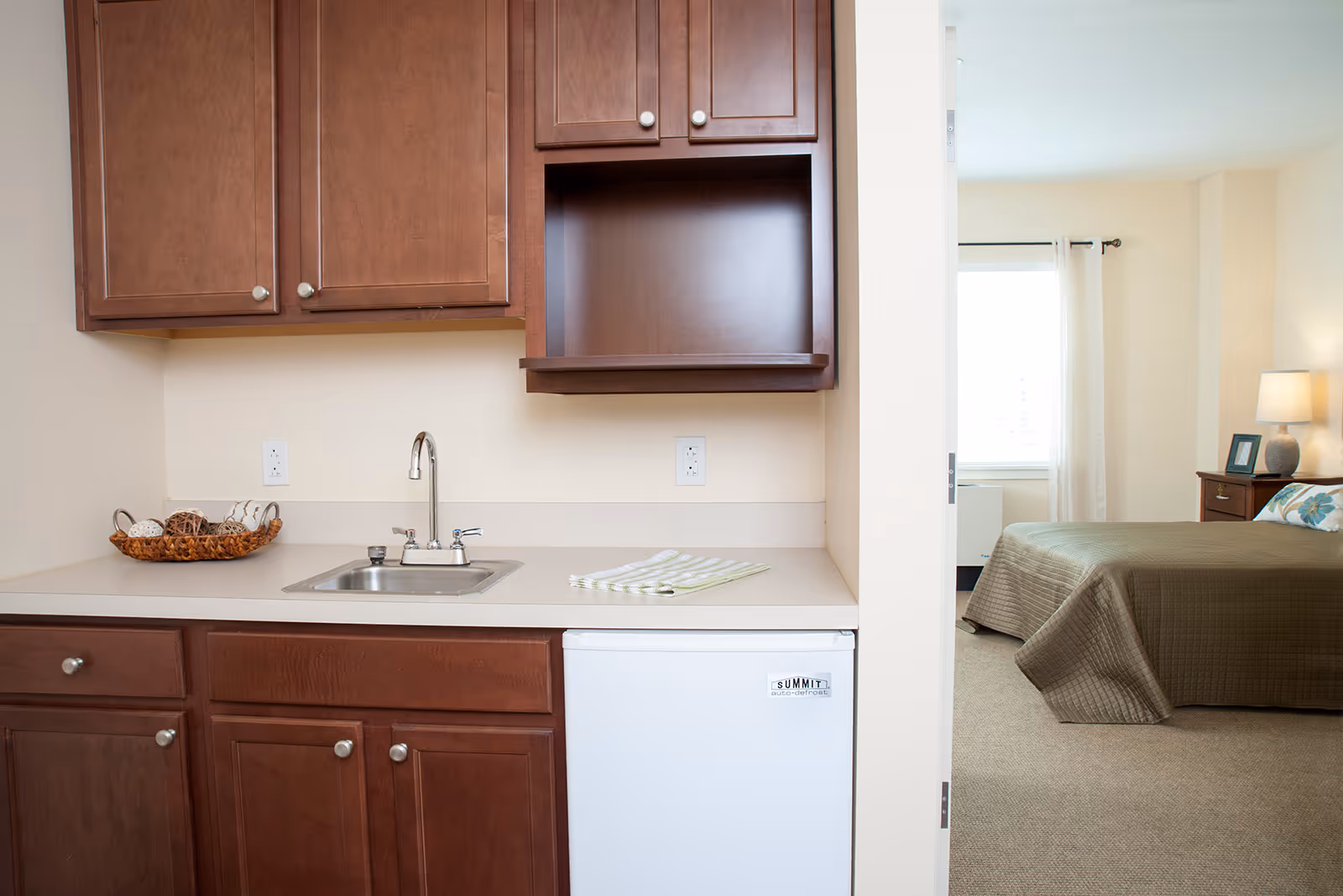 A small kitchenette with dark wooden cabinets, a stainless steel sink, and a white mini refrigerator. A basket with decorative items is on the counter next to a folded striped cloth. To the right, an open doorway leads to a bedroom with a bed covered in a brown quilt, a nightstand with a lamp, and a window with white curtains.