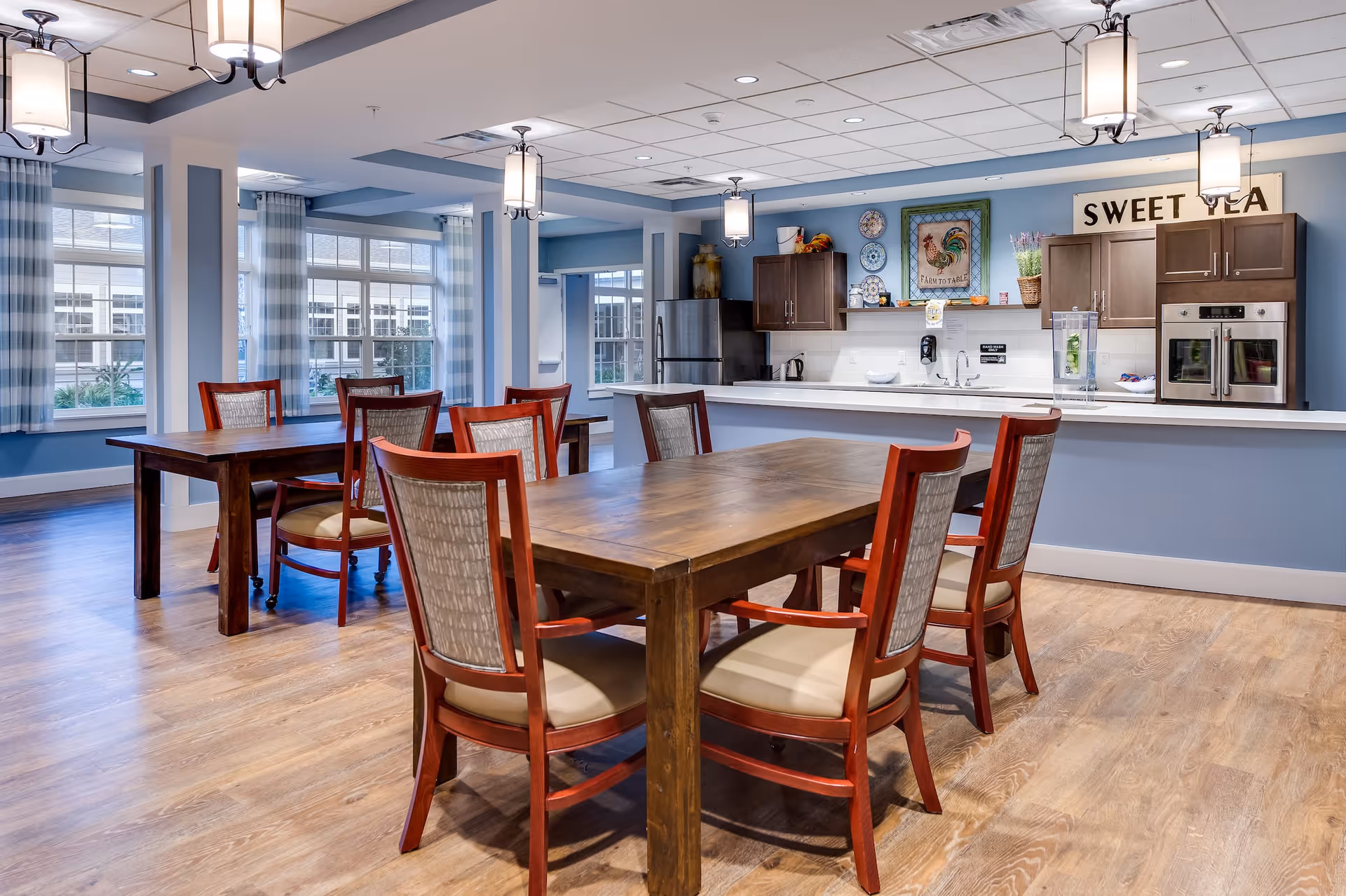 A bright dining area with wooden tables and chairs with cushioned seats. The room features large windows with blue and white checkered curtains, light wood flooring, and soft blue walls. In the background, there is a kitchen area with dark wood cabinets, stainless steel appliances, and a sign that reads 'SWEET TEA'. The kitchen counter has a water dispenser and decorative items including plates and a framed rooster picture.