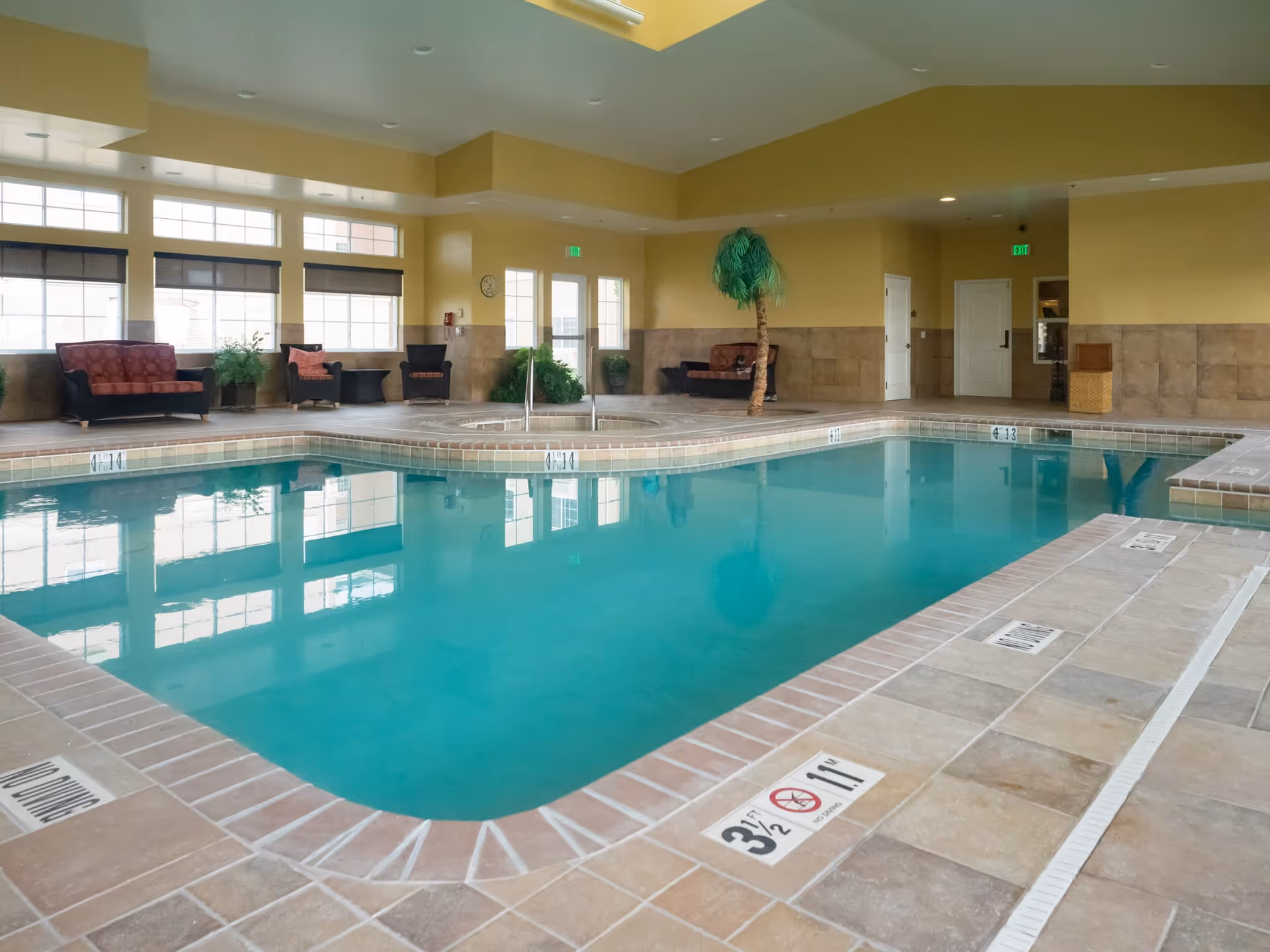 Indoor swimming pool area with clear blue water, surrounded by beige tiled flooring. Several cushioned chairs and sofas are arranged along the walls, with some green plants and a decorative palm tree near the pool. Large windows allow natural light to fill the room.