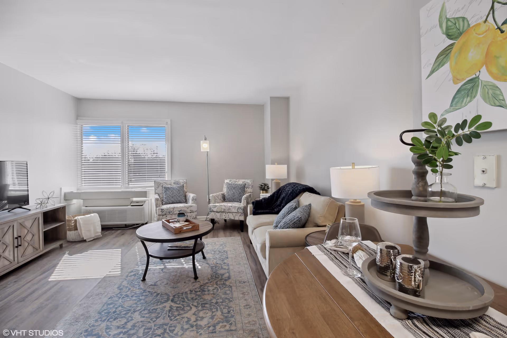 A bright and cozy living room with two patterned armchairs and a beige sofa adorned with blue and gray pillows. A round wooden coffee table sits on a blue patterned rug in the center. There is a wooden TV stand with a flat-screen TV on the left side near a window with white blinds. A small round table with decorative items and a glass of water is in the foreground on the right. The walls are painted light gray, and a painting of yellow lemons hangs on the right wall.