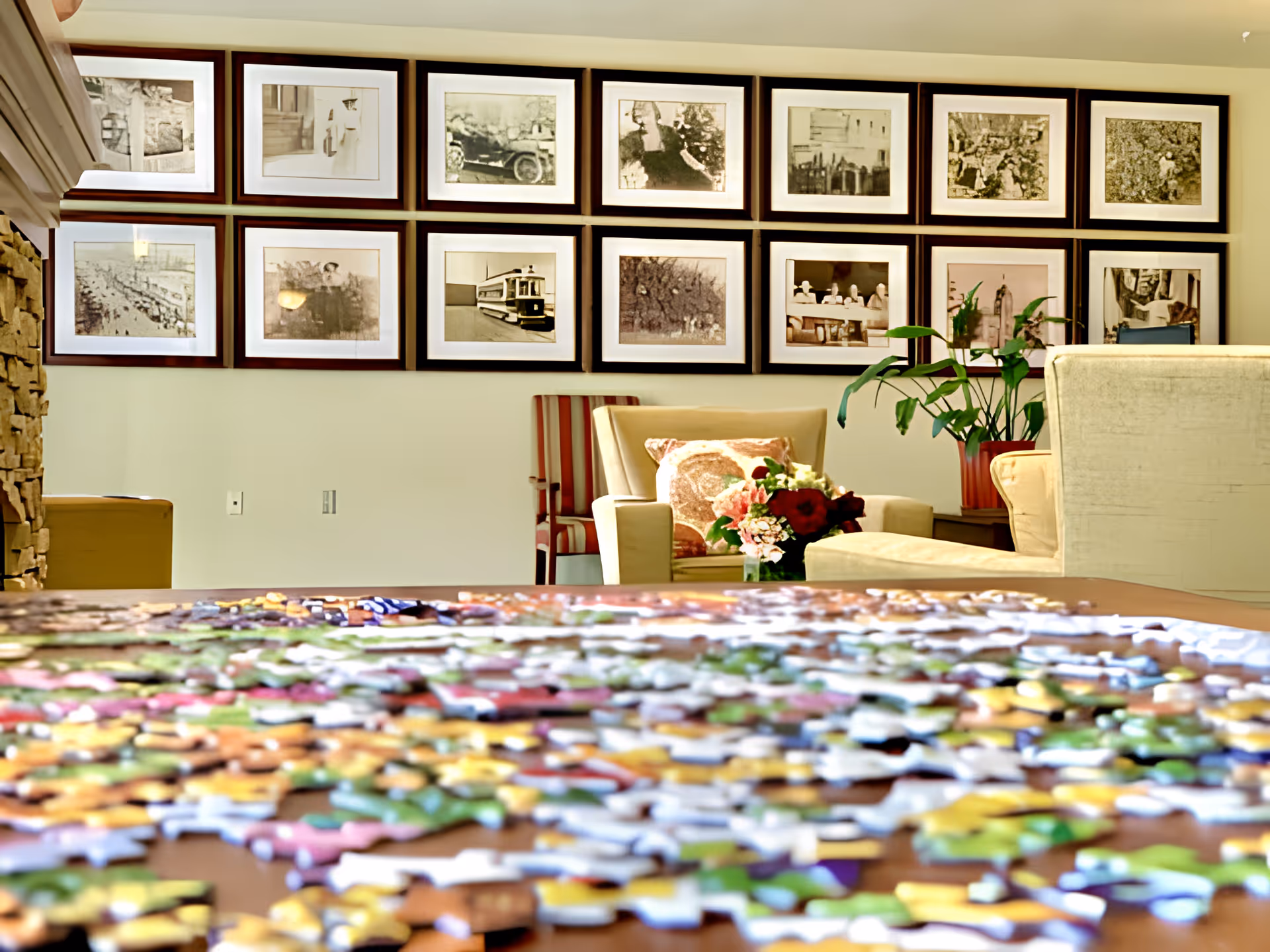 A cozy living room area with a table covered in colorful puzzle pieces in the foreground. In the background, there are beige armchairs, a striped chair, a side table with a potted plant, and a bouquet of flowers. The wall behind features a grid of framed black and white photographs.