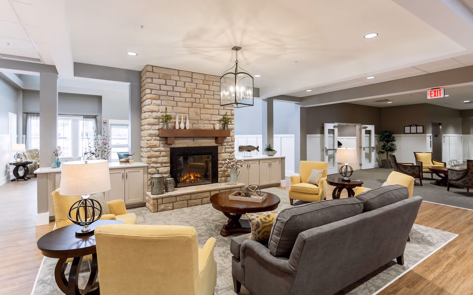 A cozy senior living facility common area with a stone fireplace as the focal point. The room features a gray sofa, yellow armchairs, wooden coffee and side tables with lamps, and decorative items on the mantle and tables. The space has a warm and inviting atmosphere with soft lighting and neutral tones.