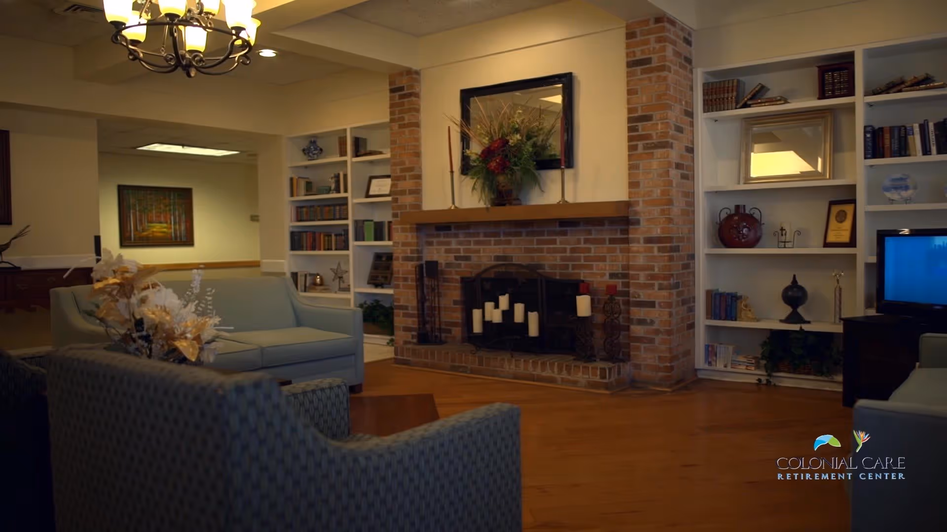 A cozy living room area with a brick fireplace decorated with candles and a floral arrangement above the mantel. There are built-in white bookshelves on either side of the fireplace filled with books and decorative items. Comfortable seating including a blue sofa and armchairs are arranged around the fireplace. A chandelier hangs from the ceiling, and a television is visible on the right side. The room has warm lighting and wooden flooring.