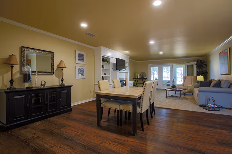 Interior view of a senior living facility showing a dining area with a table and four chairs in the foreground, and a living room with a sofa, armchair, coffee table, and built-in shelves with a TV in the background. The room has wooden flooring, beige walls, and ceiling lights.