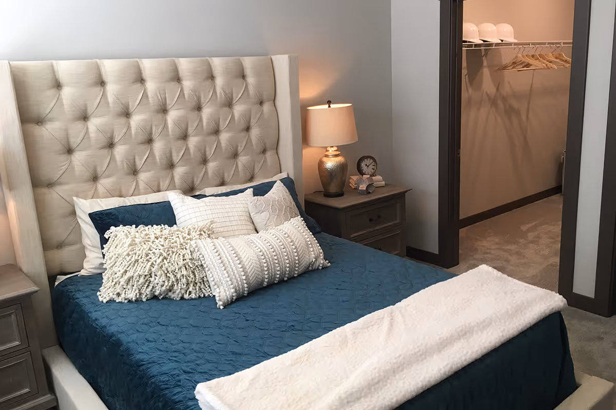 Stylish bedroom with a tufted headboard, teal bedding and decorative pillows, a nightstand with lamp, and an open walk-in closet.
