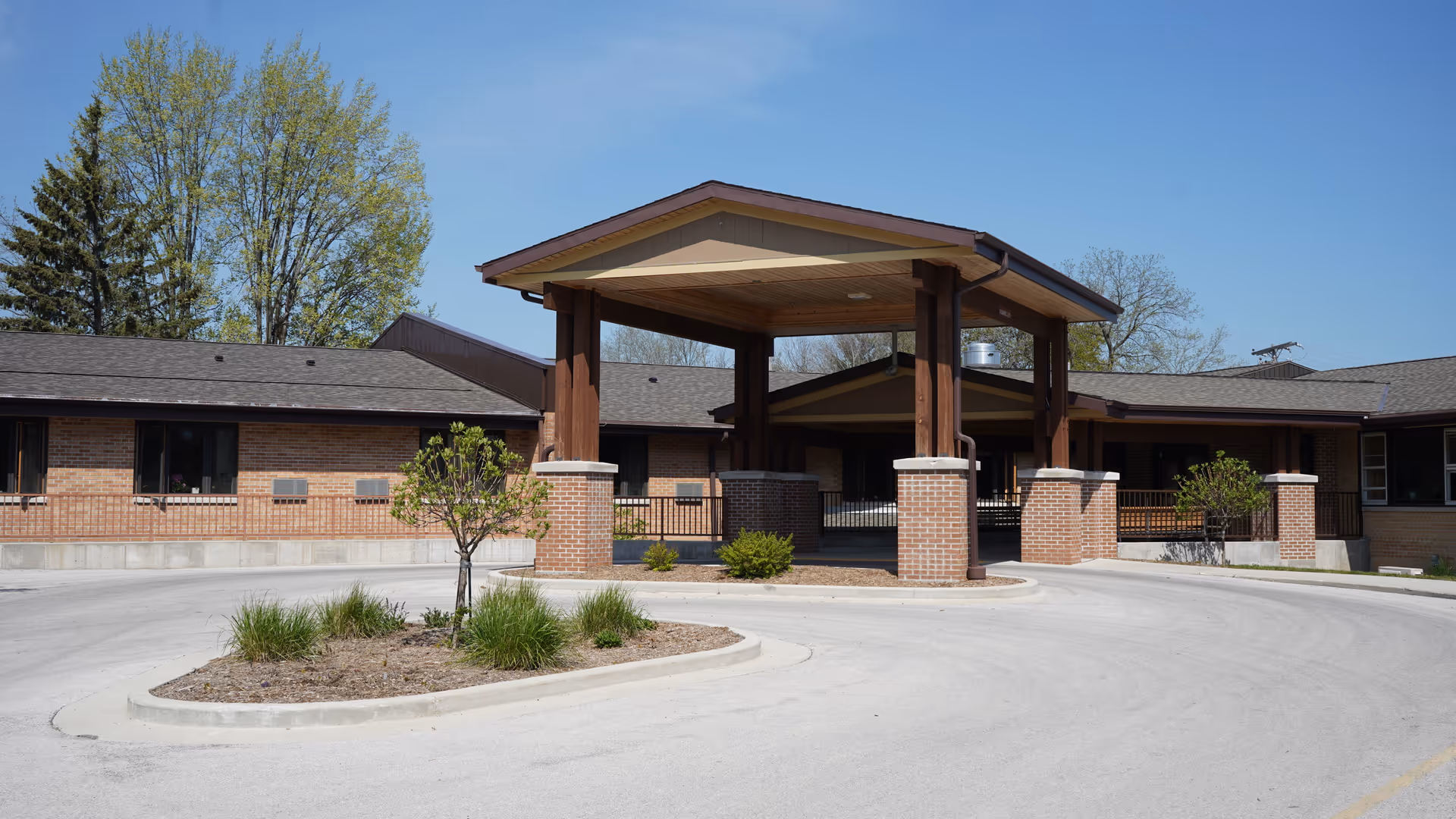 Front exterior view of Pine Haven Christian Communities Giddings Avenue Campus showing a covered entrance with wooden beams and brick pillars, surrounded by a circular driveway and some landscaping with small trees and bushes under a clear blue sky.