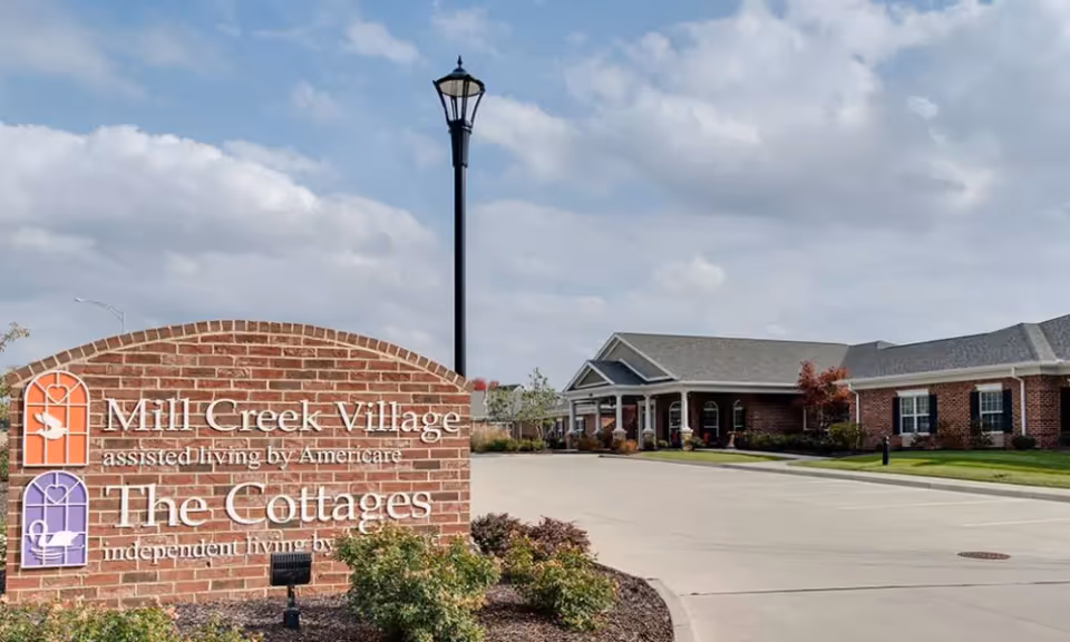 Entrance sign for Mill Creek Village next to a single-story brick senior living building, driveway, lamp post, and cloudy sky.