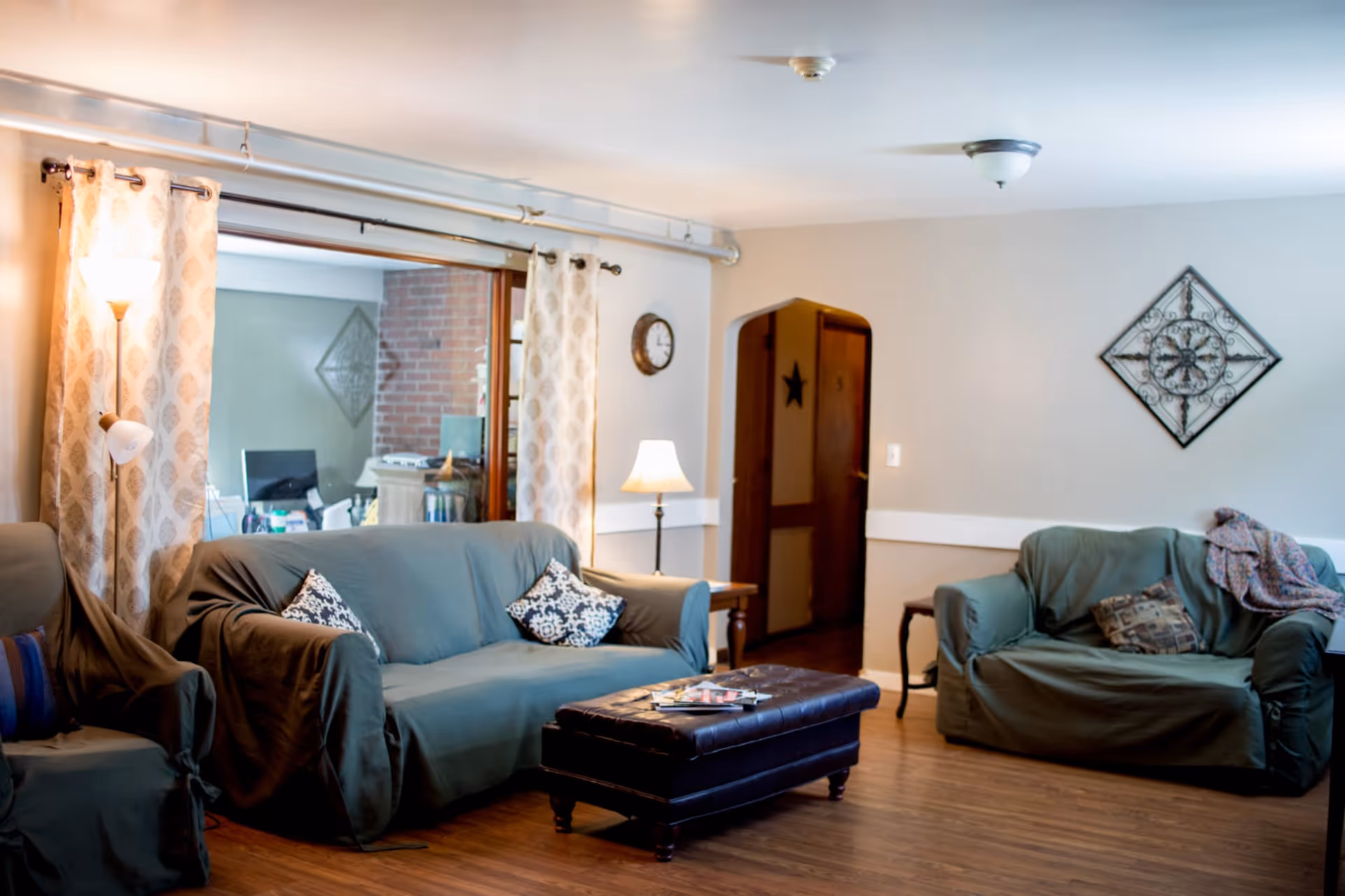 A cozy living room with two green-covered sofas, a dark brown leather ottoman with magazines on top, a floor lamp, a table lamp on a side table, patterned curtains, a wall clock, and decorative wall art. The room has wooden flooring and a doorway leading to another area.
