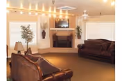 A cozy common living room with two leather sofas facing a fireplace with a wall-mounted TV, windows, lamps, plants, and ceiling string lights.