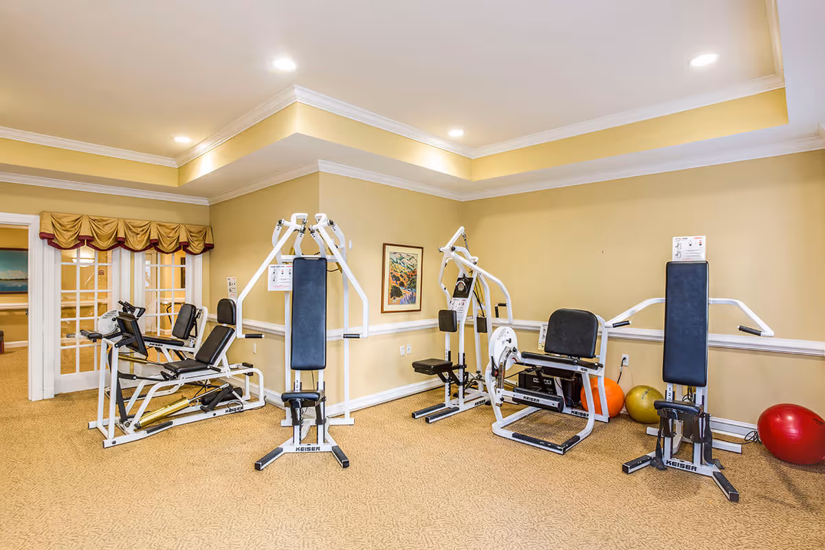 A small gym room with beige walls and carpeted floor, equipped with several white exercise machines and colorful exercise balls in the corner. The room has recessed lighting and a framed picture on the wall.
