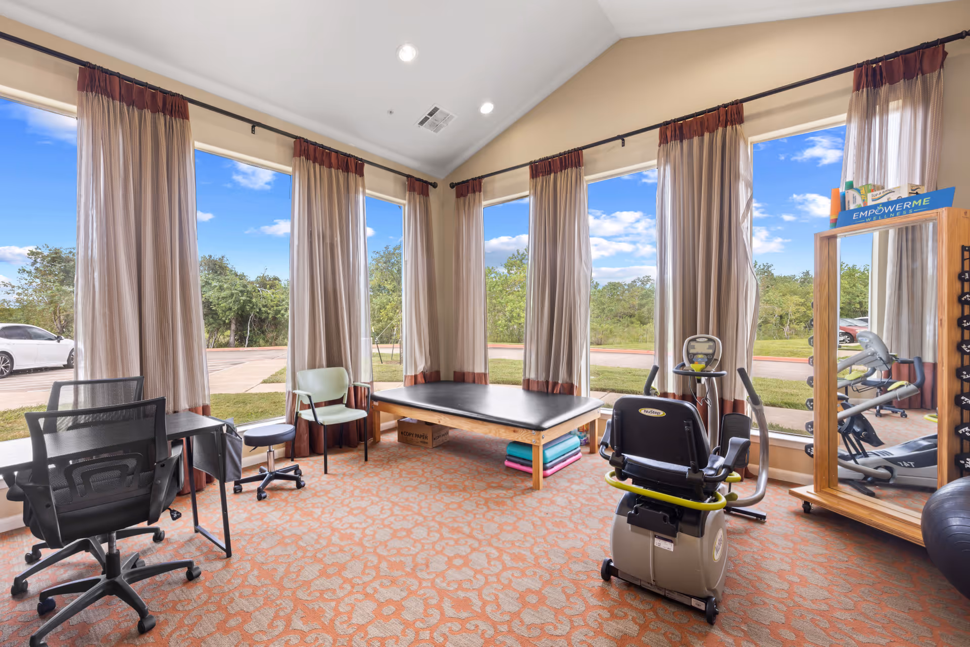 A bright room with large windows covered by sheer curtains, featuring exercise equipment including a recumbent bike, a padded therapy table, chairs, a desk with office chairs, and a large mirror with weights beside it. The room has a patterned carpet and overlooks a green outdoor area with trees and a parking lot.