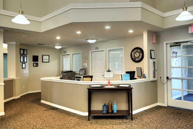 Reception area with a curved front desk, a small table with decorative vases, a clock on the wall, framed pictures, and a glass door entrance.
