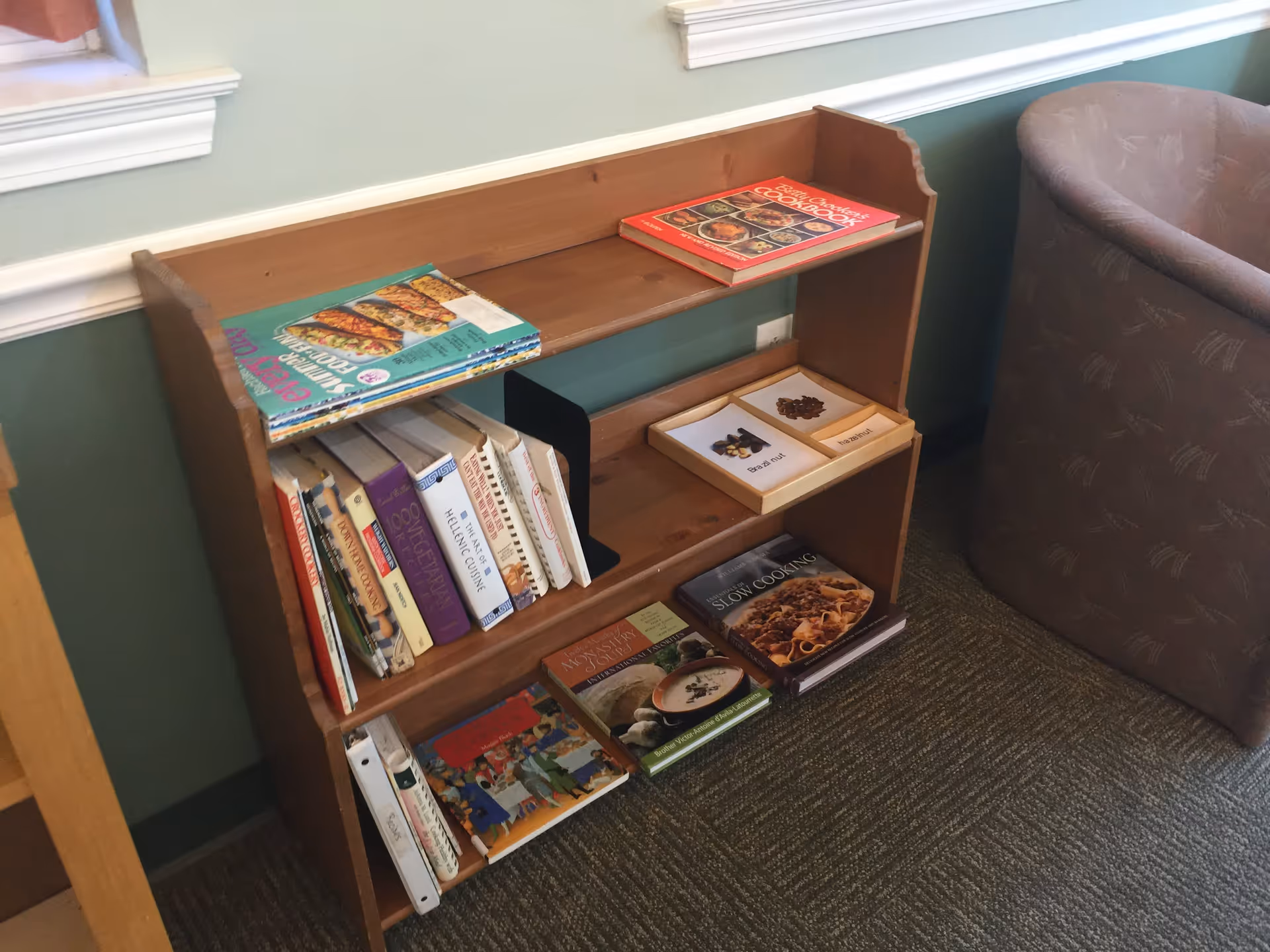 Small wooden bookshelf filled with cookbooks and magazines next to a padded armchair in a room.