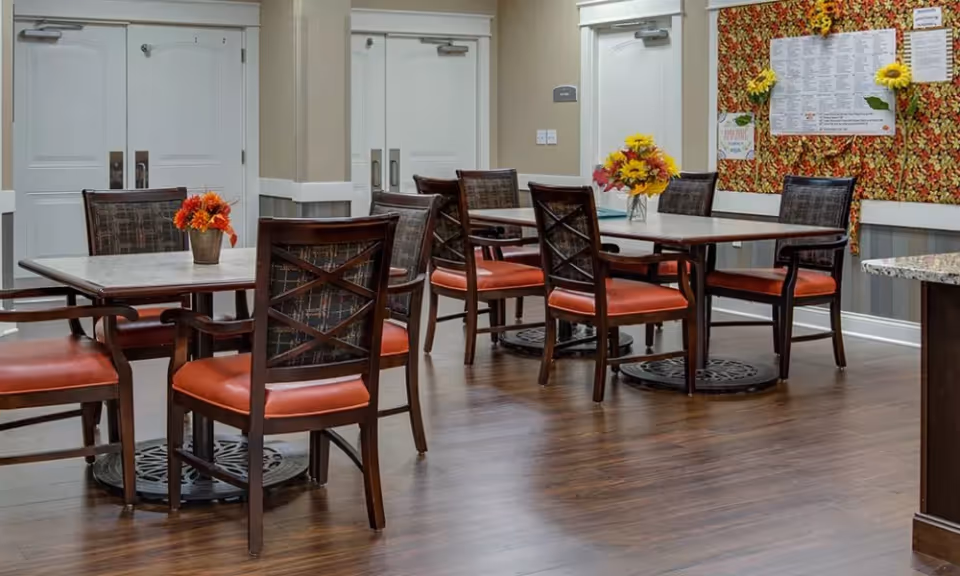 A dining area with multiple tables and chairs, small floral centerpieces, and a decorative bulletin board on the wall.