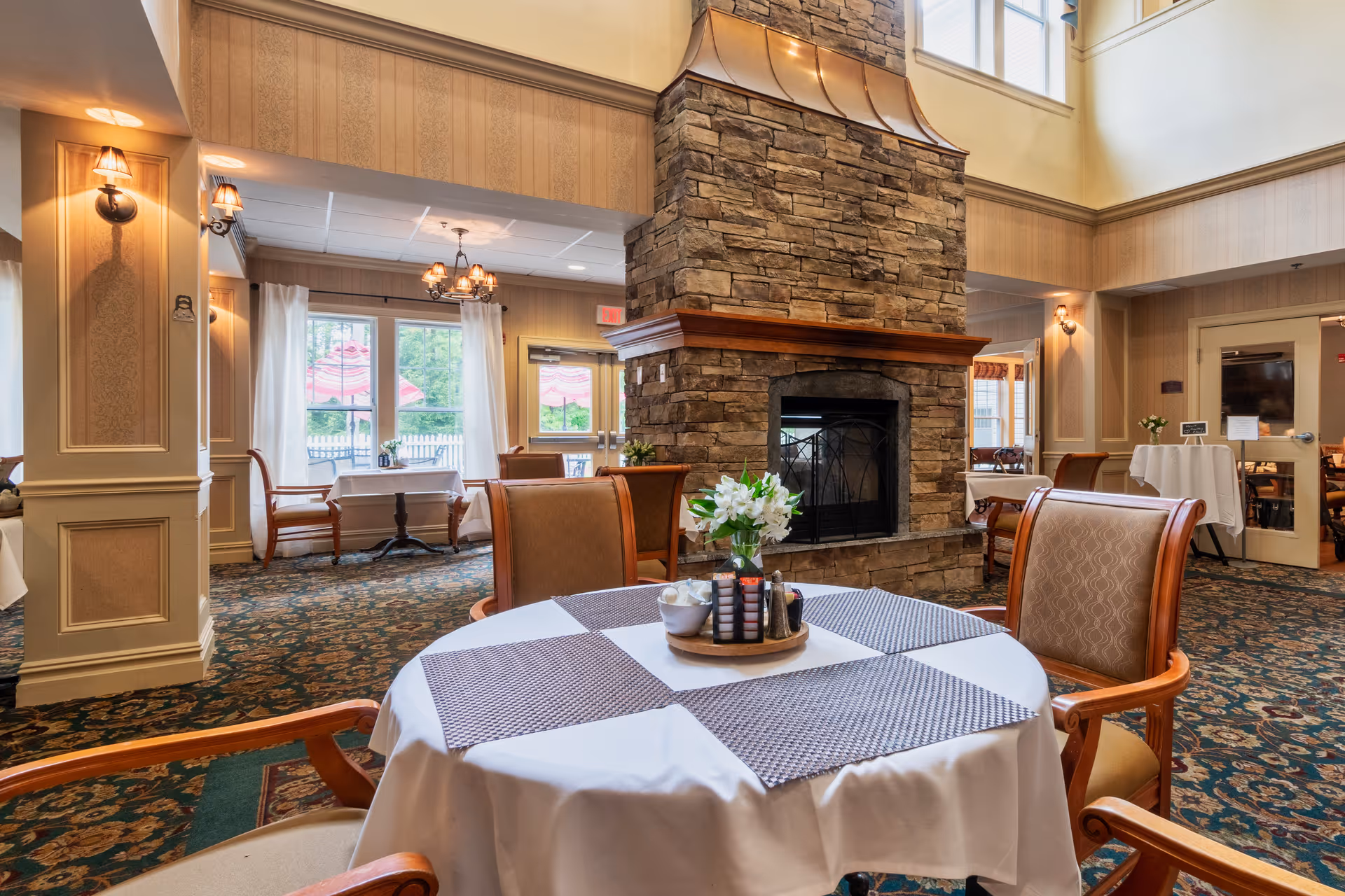 Dining room with a round table set with placemats and flowers, upholstered chairs, and a central stone fireplace.