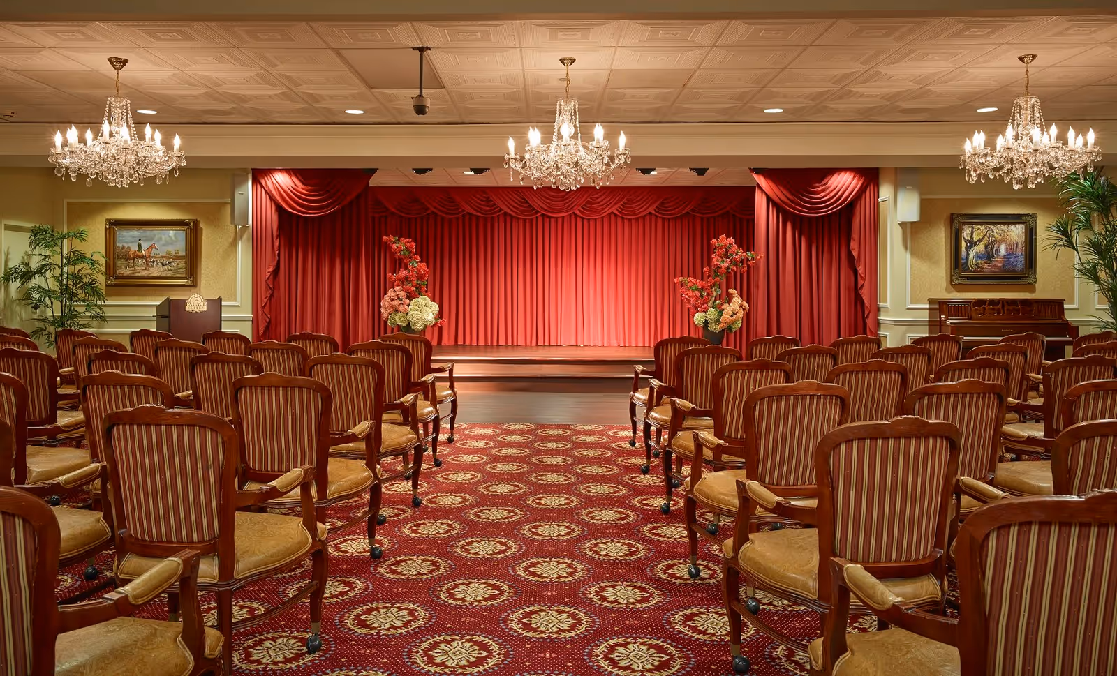 An elegant auditorium with rows of wooden chairs featuring striped upholstery facing a stage with red curtains. The room is decorated with chandeliers, floral arrangements on the stage, framed paintings on the walls, and potted plants.