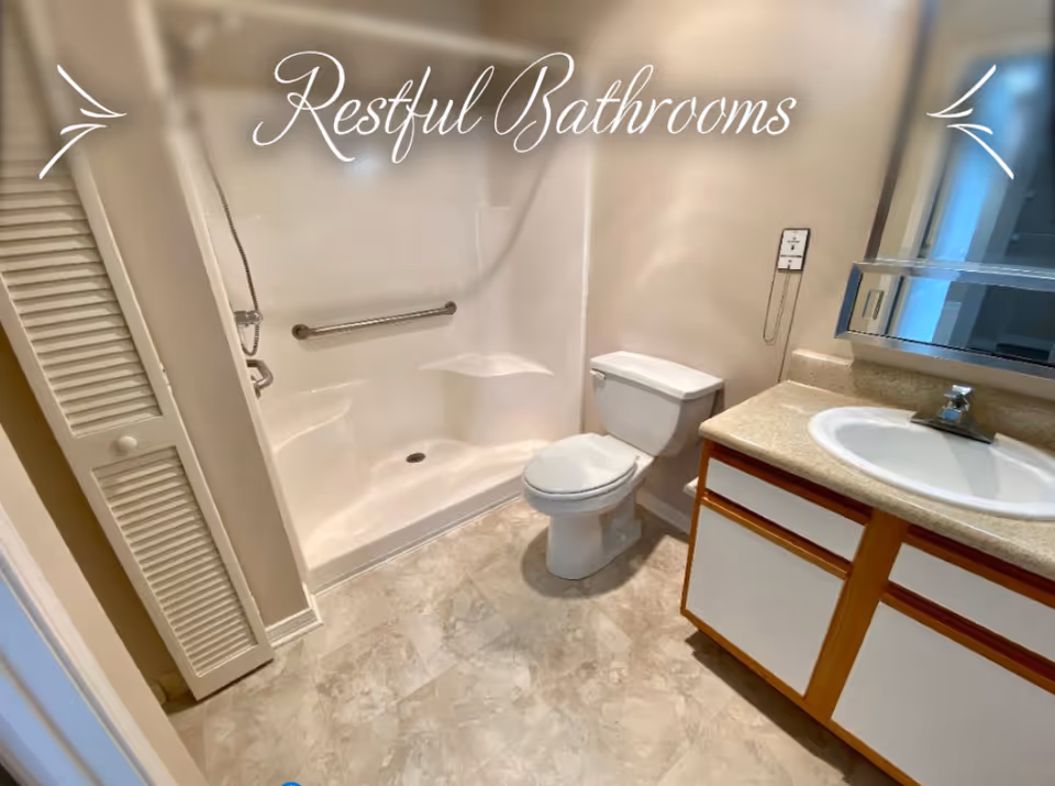 A clean and accessible bathroom featuring a walk-in shower with a built-in seat and grab bar, a white toilet, and a vanity with a sink and mirror. The floor has a light-colored tile, and there is a louvered door to the left.