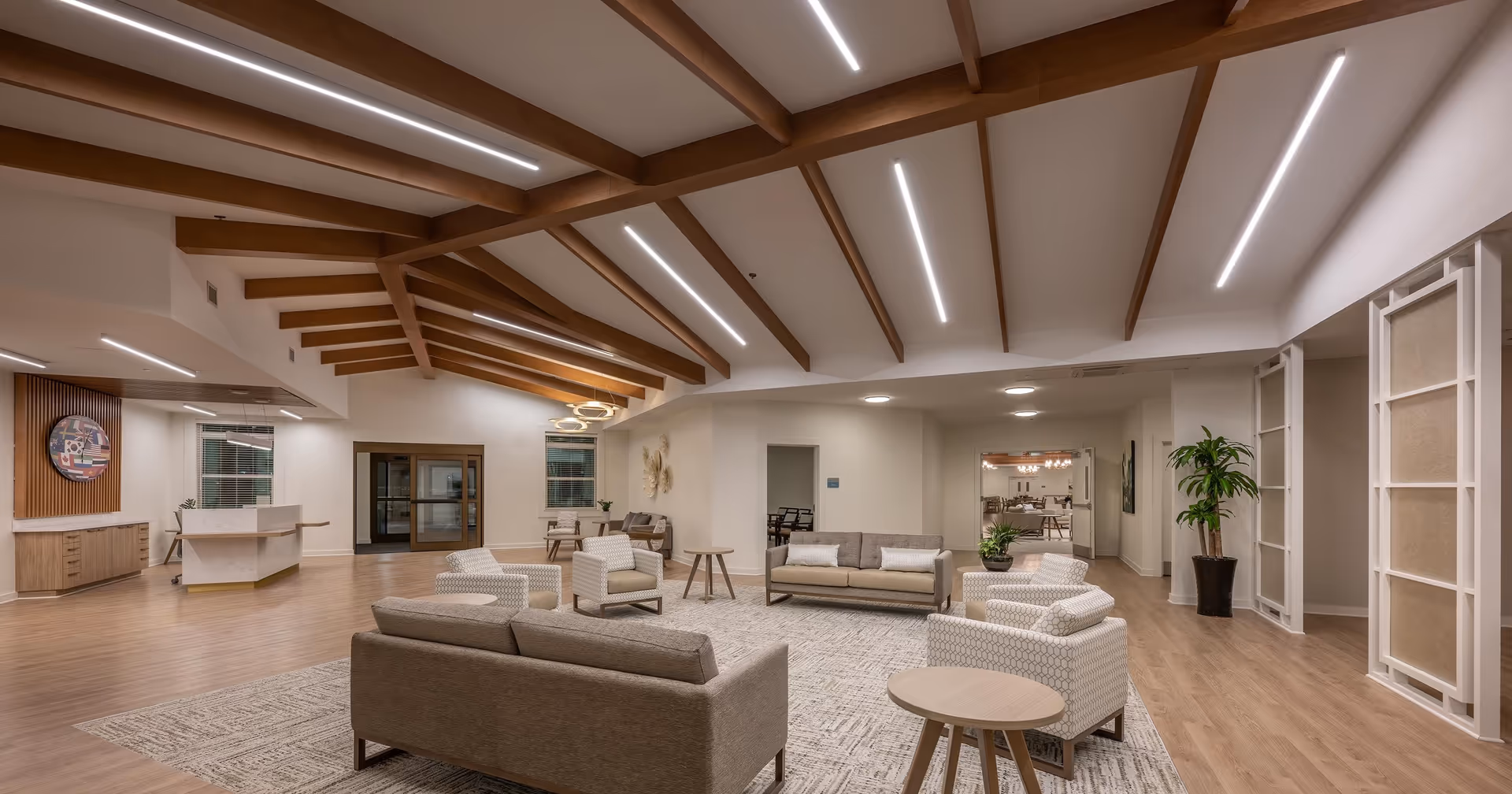 A spacious and modern senior living facility common area with wooden ceiling beams, recessed lighting, beige sofas and armchairs arranged around a large rug, small round wooden tables, and a reception desk in the background. The room has light wood flooring and neutral-colored walls with decorative elements and plants.