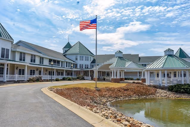 Exterior view of Oak Grove Christian Retirement Village showing a large building with green metal roofs, white siding, and multiple windows. There is a circular driveway with an American flag on a flagpole in the center, surrounded by landscaping with rocks and a small pond. The sky is partly cloudy.