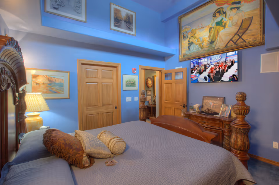 Decorated bedroom with a large wooden bed, blue walls, framed artwork, wooden doors, and a TV mounted on the wall.
