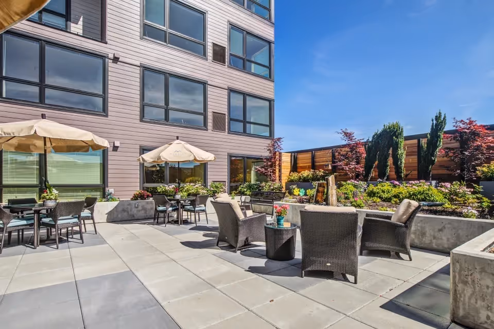 Outdoor patio area at Merrill Gardens at Renton Centre with several wicker chairs and tables, some shaded by beige umbrellas. The patio is surrounded by a garden with various plants and small trees, and the building's exterior with large windows is visible in the background under a clear blue sky.