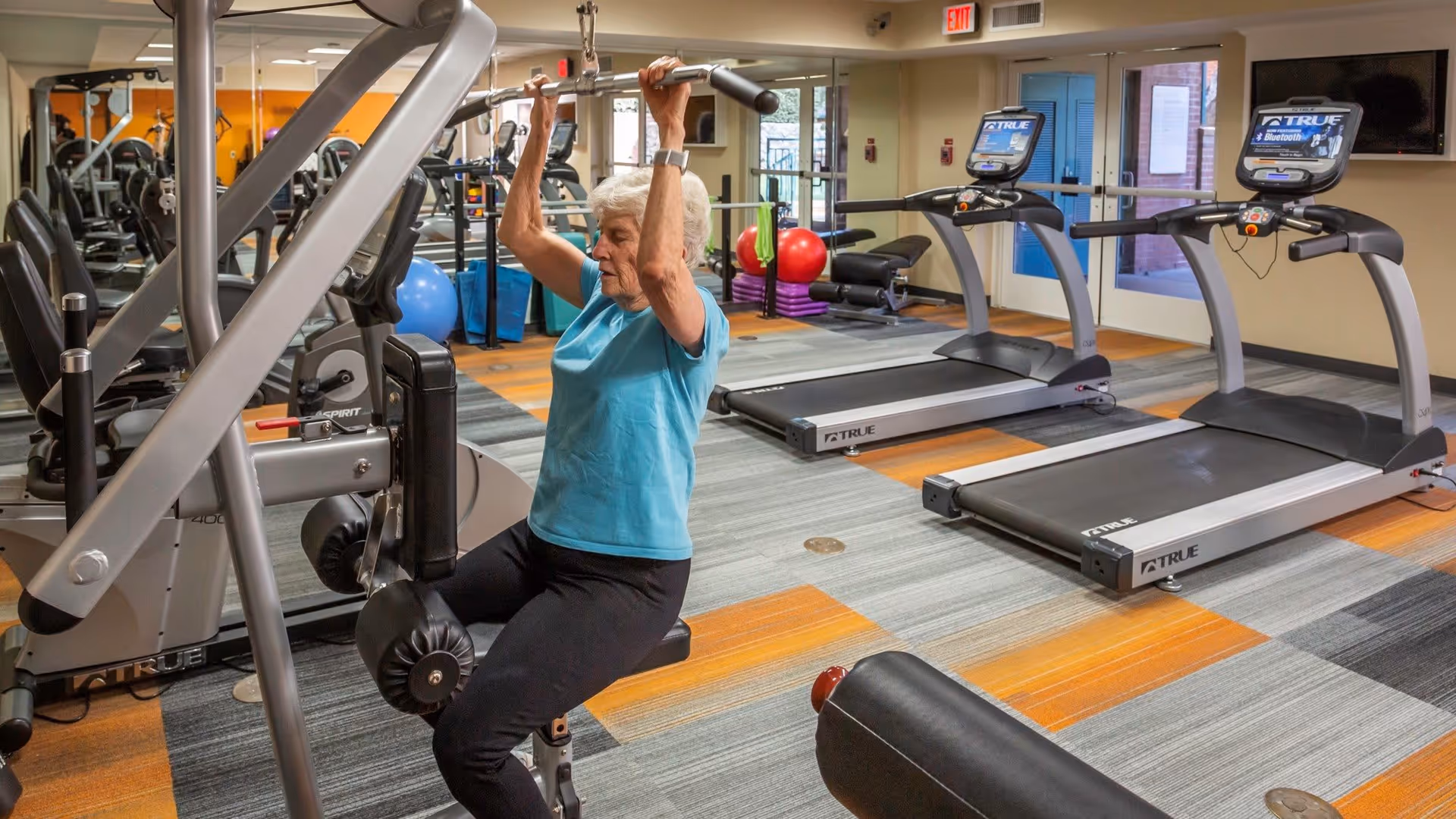An older woman using a lat-pulldown machine in a fitness room with treadmills and other exercise equipment.