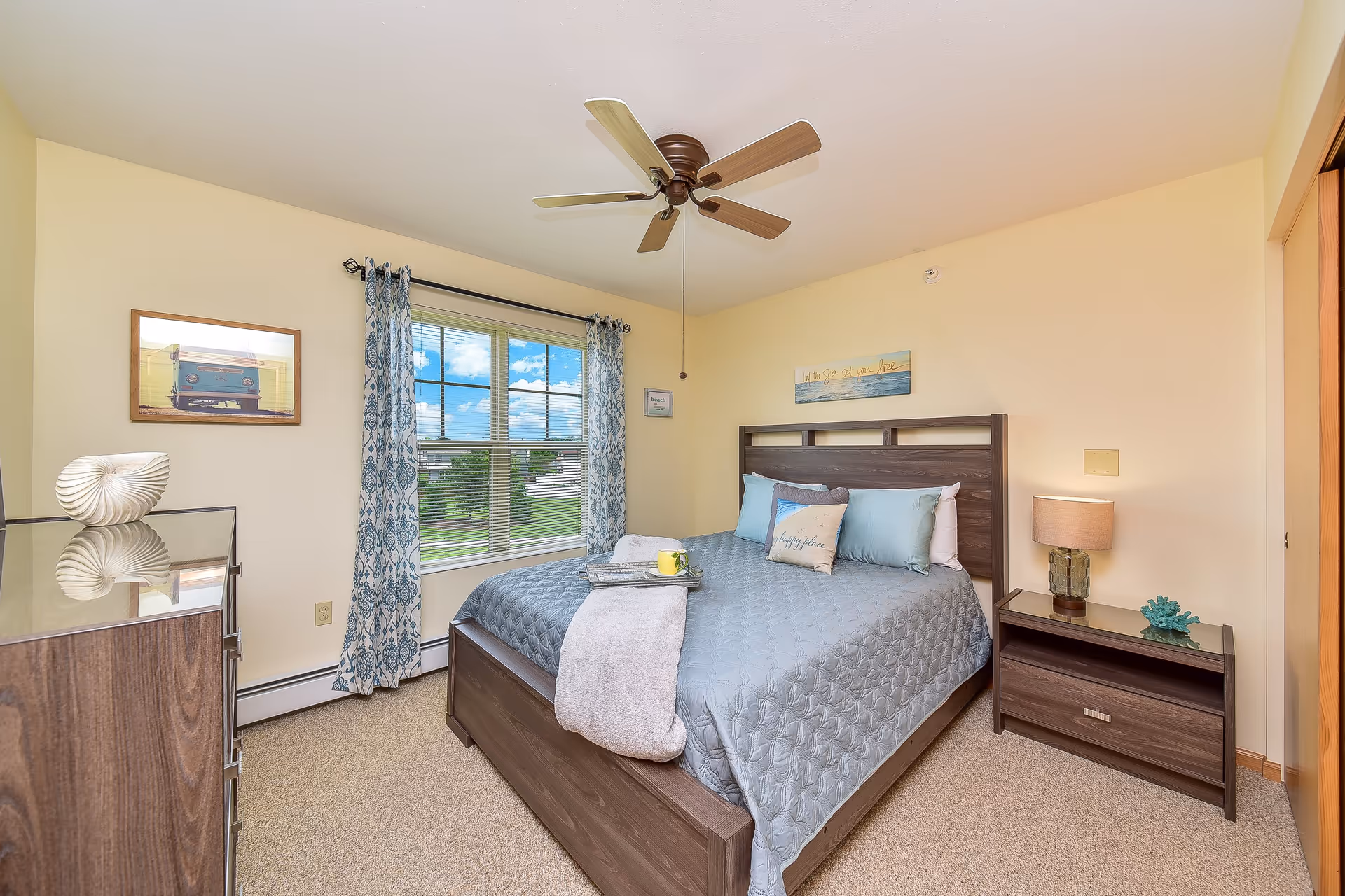 A bright bedroom with a large window covered by blue patterned curtains. The room features a wooden bed with a gray quilted bedspread, several pillows, and a folded blanket with a tray holding a yellow cup. There is a wooden nightstand with a lamp and decorative items, a matching dresser with a white shell decoration, and a ceiling fan overhead. The walls are painted light yellow and decorated with framed pictures.
