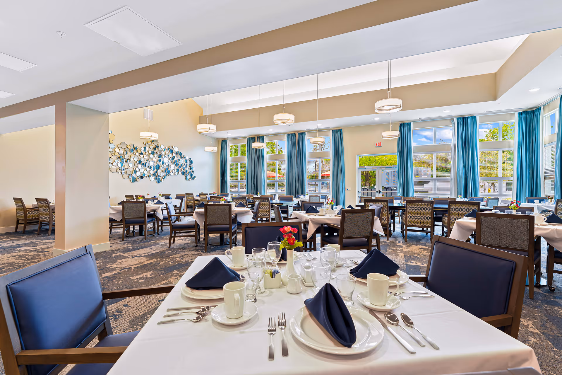Spacious dining room with neatly set tables, blue curtains, and large windows letting in daylight.