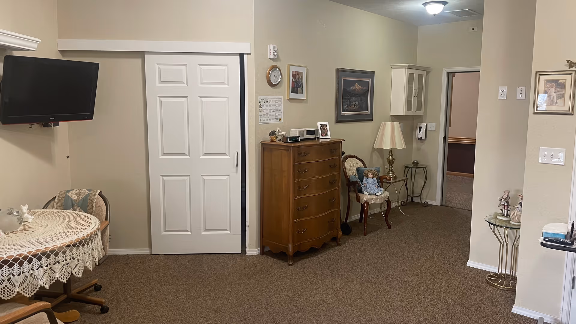 Interior view of a room in a senior living facility featuring a small round table with a lace tablecloth and two chairs, a wall-mounted TV, a wooden chest of drawers with a clock and framed photos on top, a decorative chair with a doll seated on it, a side table with a lamp, and framed artwork on the walls. The room has beige walls and carpeted floor.