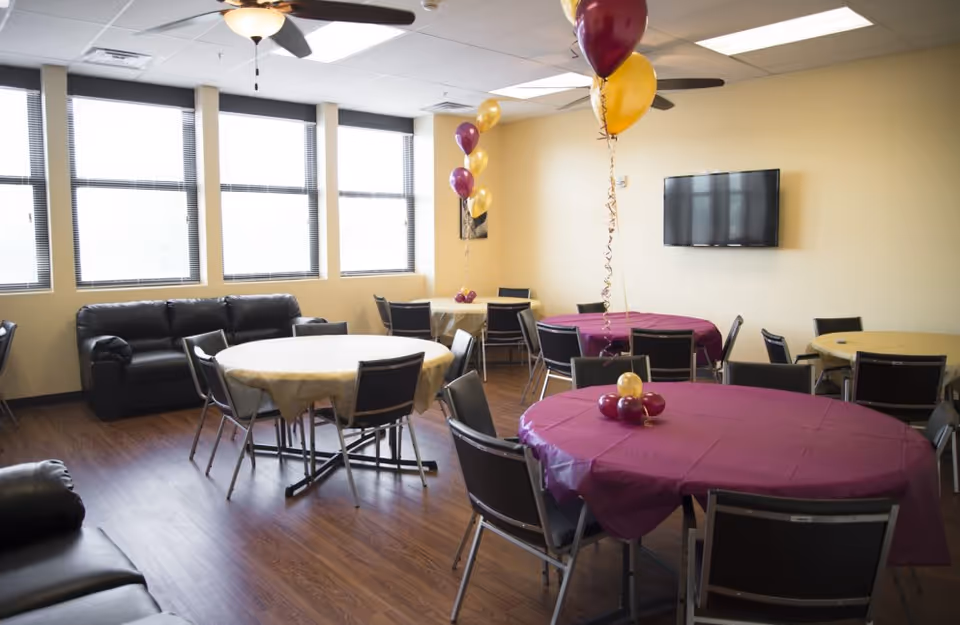 A bright common room with round tables covered in purple and yellow tablecloths, balloons, chairs, a couch and a wall-mounted TV.