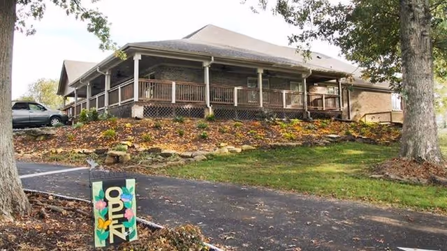 Exterior view of a single-story building with a large covered porch and wooden railings, surrounded by a landscaped garden with trees and a paved driveway. A colorful sign with the word 'OPEN' is placed near the driveway.