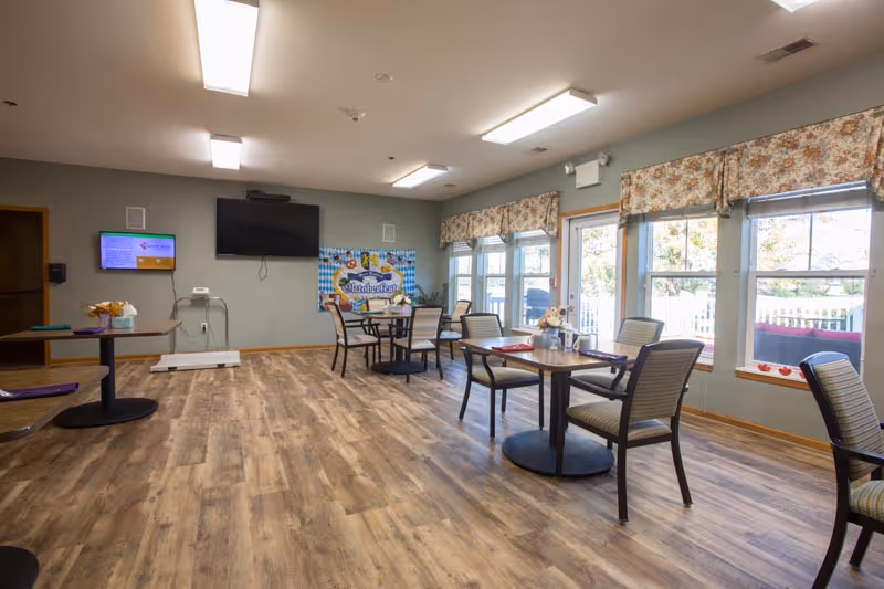 A bright dining room in an assisted living facility with wooden floors, several round tables with chairs, large windows with floral valances letting in natural light, a wall-mounted TV, and a colorful Oktoberfest banner on the far wall.
