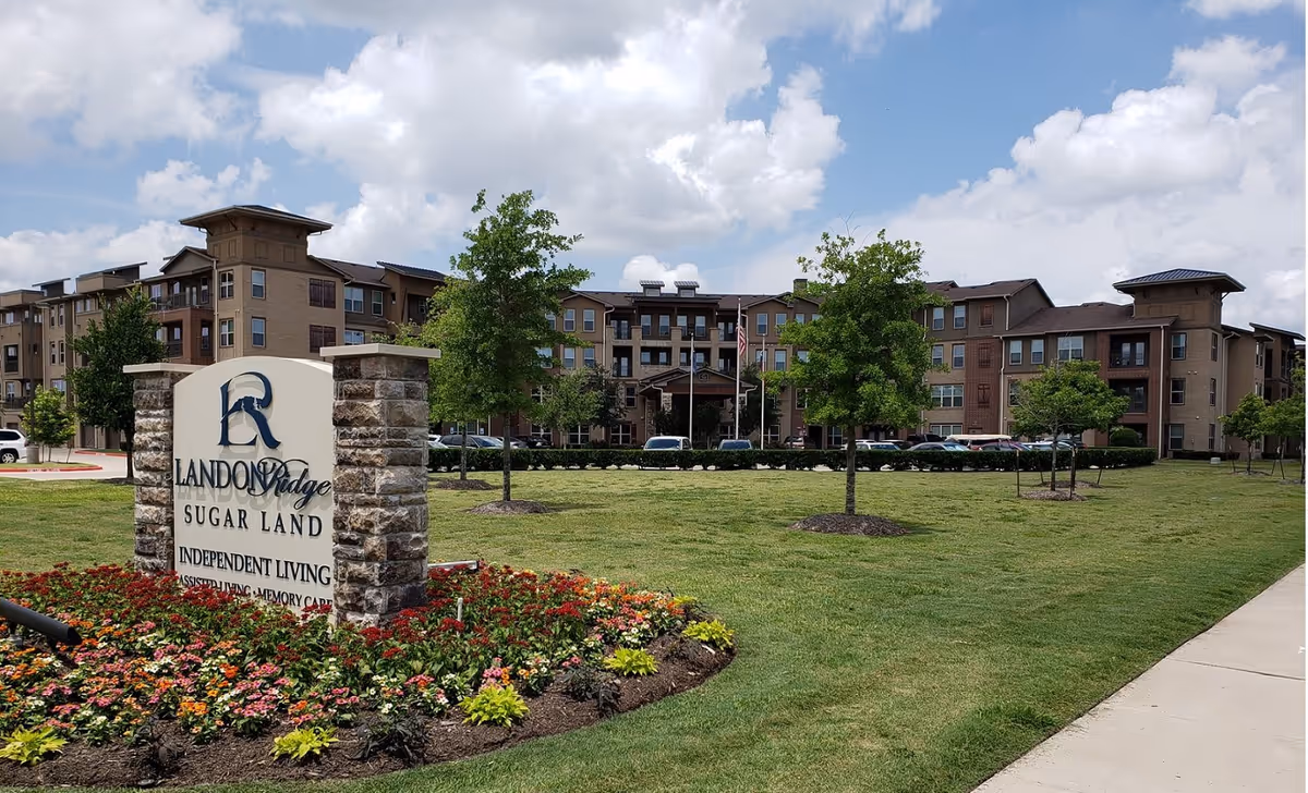 Exterior view of Landon Ridge Sugar Land Assisted Living & Memory Care facility with a large green lawn, trees, and a stone sign surrounded by colorful flowers in the foreground. The multi-story building is visible in the background under a partly cloudy sky.