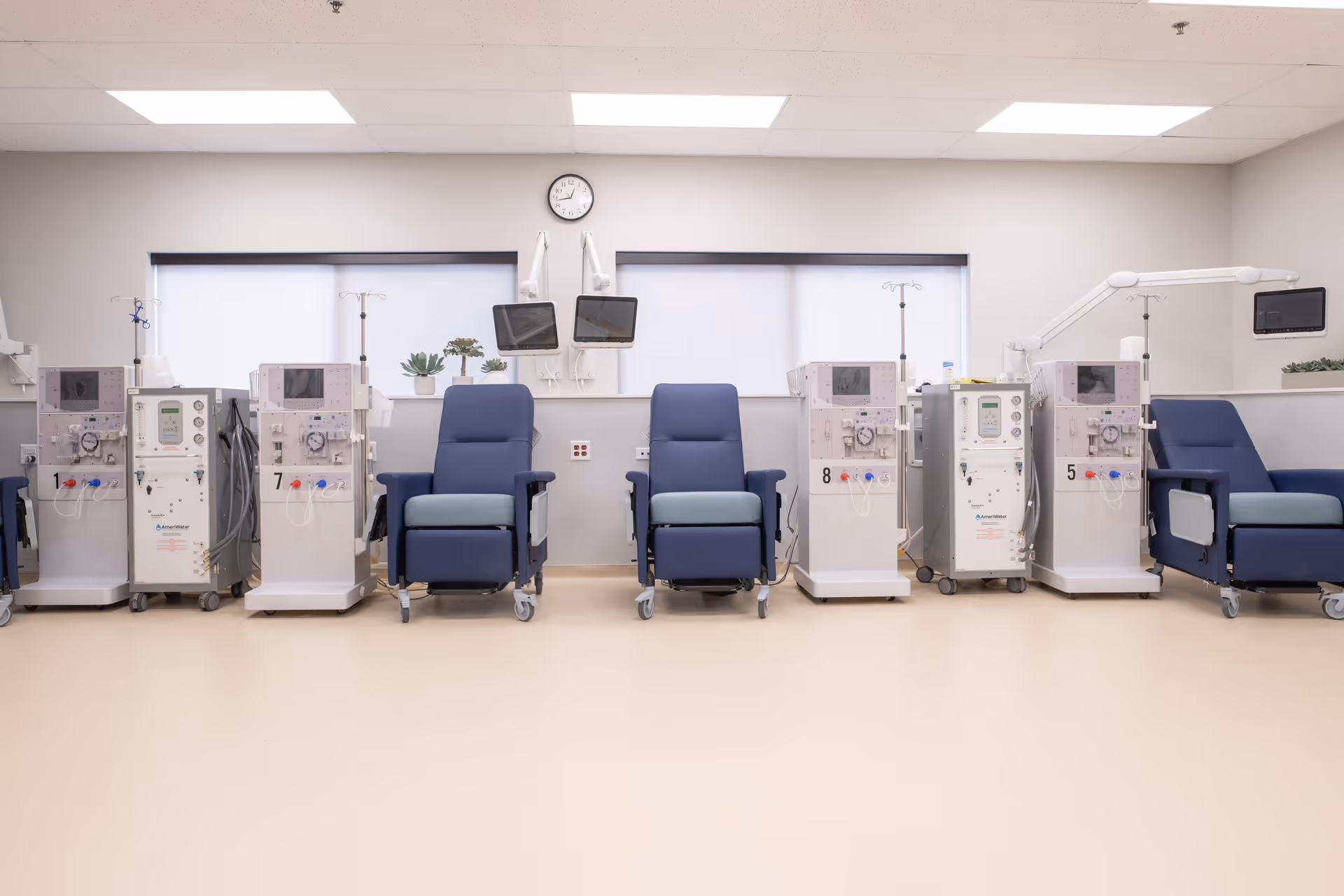 A clean and well-lit medical treatment room with multiple dialysis machines lined up against the wall, each paired with a comfortable blue recliner chair. The room has large windows with blinds, two wall-mounted monitors, a clock, and some small potted plants on the windowsill.