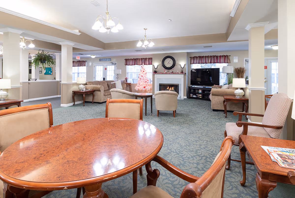 A spacious living room area in a senior living facility with beige walls and carpeted floor. The room features multiple seating options including sofas, armchairs, and wooden chairs around a polished wooden table. A fireplace with a clock above it is lit, and a pink Christmas tree is placed nearby. There is a large flat-screen TV on a stand to the right, and several lamps and decorative plants are arranged throughout the room. The room is well-lit with ceiling lights and natural light from windows with red valances.