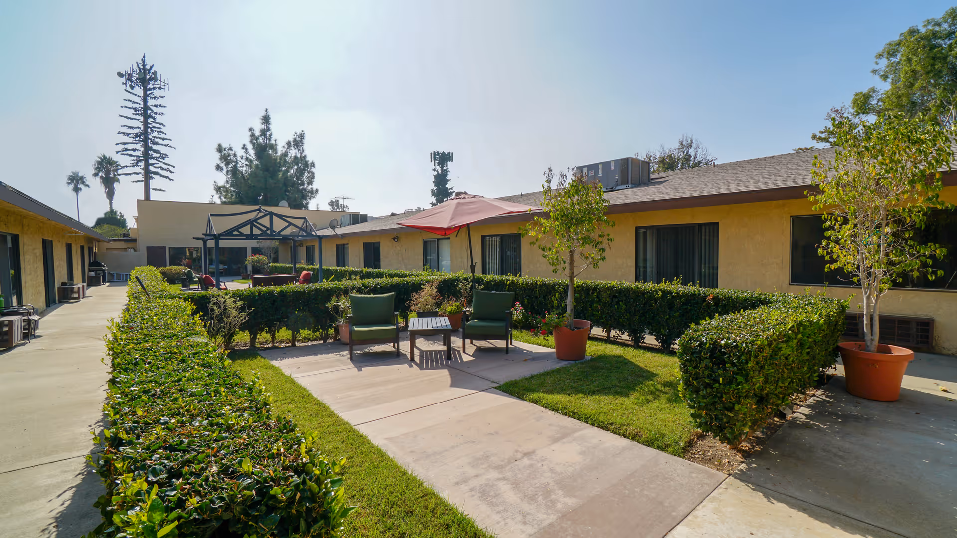Outdoor courtyard area at Santa Anita Assisted Living featuring a paved walkway, green hedges, two green cushioned chairs with a small table and a red umbrella, potted plants, and a pergola structure in the background. The building surrounds the courtyard with windows and doors visible.