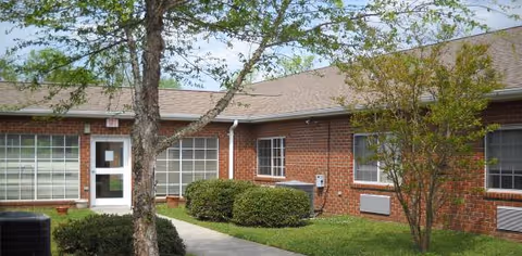 Brick, single-story senior living facility exterior with glass door, windows, trees, and shrubs.
