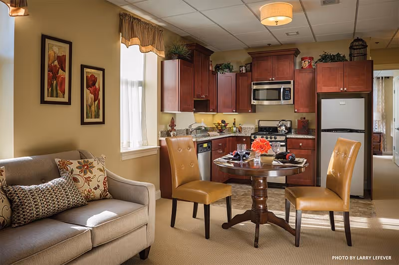 Cozy senior apartment interior showing a small kitchen, round dining table with two chairs, and a sofa by a window.