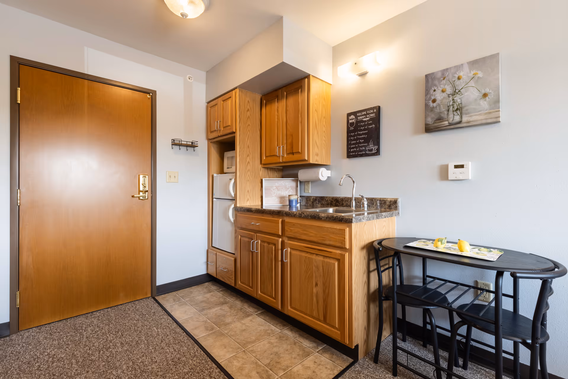 Small kitchenette area with wooden cabinets, a countertop with a sink, a microwave, and a mini refrigerator. To the right, there is a small black table with two matching chairs and a decorative placemat with lemons. The walls are light-colored with two pieces of wall art, one featuring flowers in a vase and the other a framed quote. A wooden door is visible on the left side of the image.