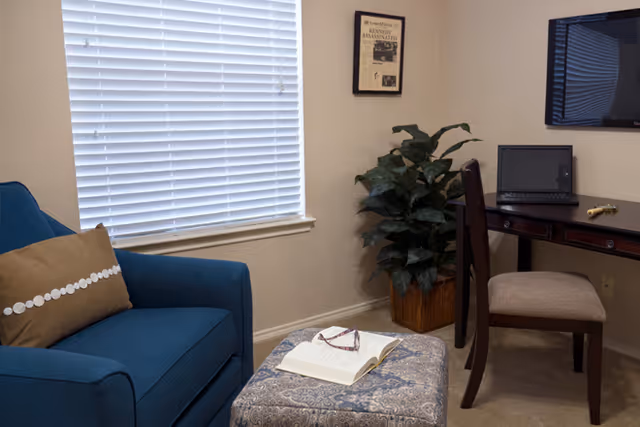A cozy room with a blue armchair, a patterned ottoman with an open book and reading glasses on top, a wooden desk with a laptop and a chair, a potted plant in the corner, a window with closed blinds, and a framed newspaper clipping on the wall.