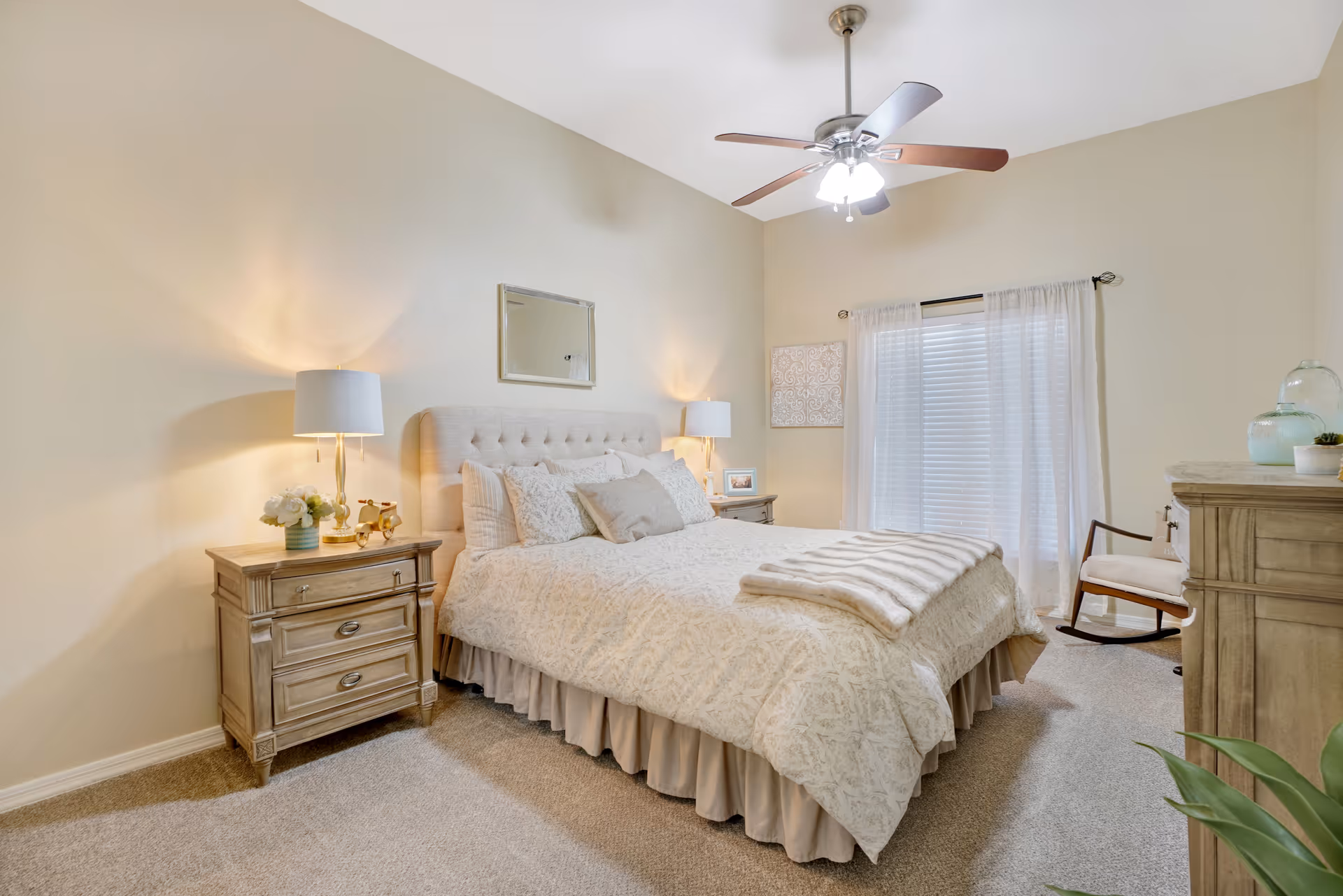 A cozy bedroom with a large bed covered in a beige patterned comforter and multiple pillows. There are two wooden nightstands on either side of the bed, each with a lamp and decorative items. A ceiling fan with lights hangs above. A window with sheer white curtains is on the far wall, next to a wooden rocking chair. A wooden dresser with decorative glass items is on the right side of the room, and a green plant is partially visible in the foreground.