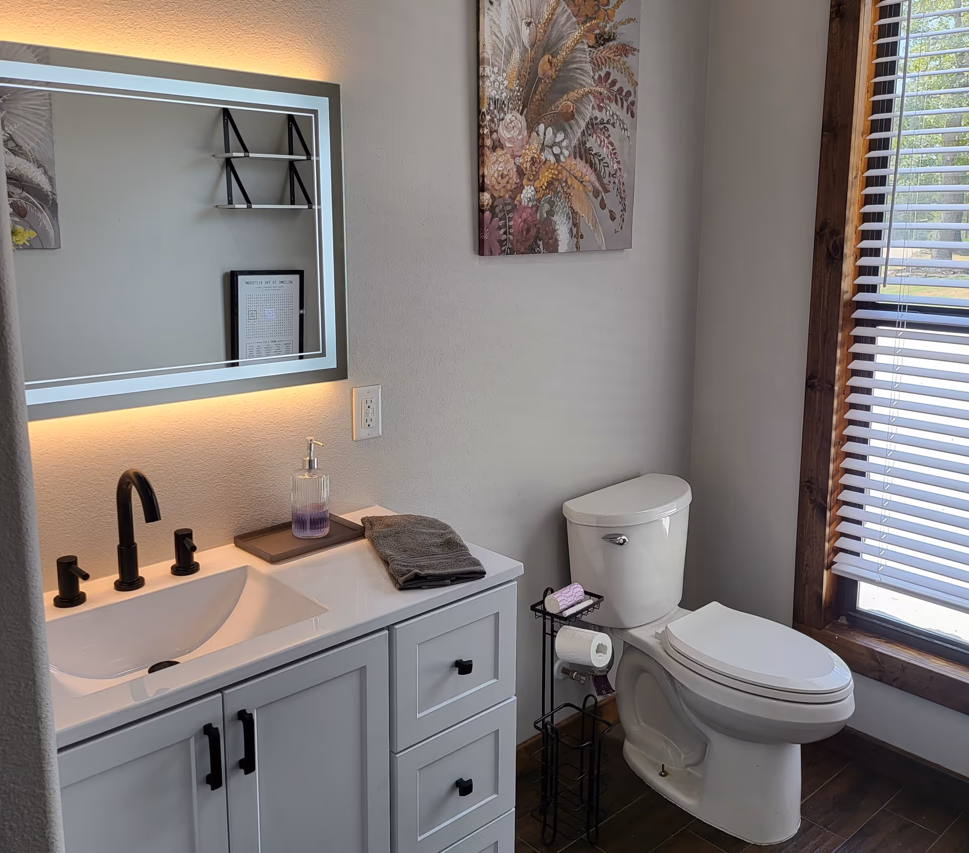A modern bathroom with a white vanity featuring black handles, a black faucet, a soap dispenser, and a folded gray towel on the countertop. Above the vanity is a rectangular mirror with built-in lighting. To the right is a white toilet next to a window with wooden trim and white blinds. A small black metal stand holds a roll of toilet paper and a pink toilet paper roll. A floral painting hangs on the wall above the toilet.