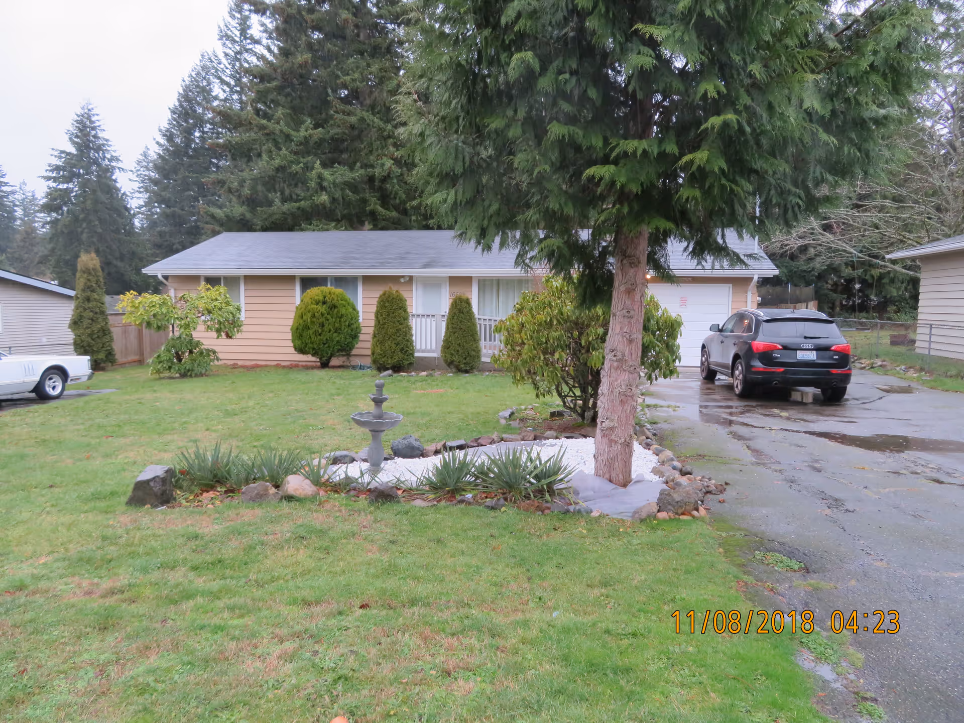 Single-story beige house with a gray roof surrounded by green grass and trees. There is a small garden with a fountain in front of the house, a driveway with a black SUV parked on it, and another white vehicle parked to the left. The scene is outdoors with tall evergreen trees in the background.