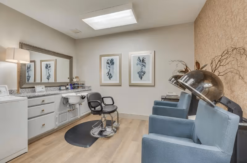 Interior view of a hair salon area in a senior living facility featuring a black salon chair in front of a large mirror and countertop with hair products. To the right, there are three blue salon chairs with hair dryers attached. The room has light wood flooring, two framed botanical prints on the wall, and a lamp on the countertop.