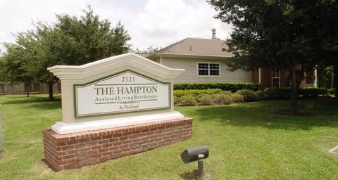 A large sign in front of a single-story building with a lawn and trees. The sign reads '2121 THE HAMPTON Assisted Living Residences At Pearland'.