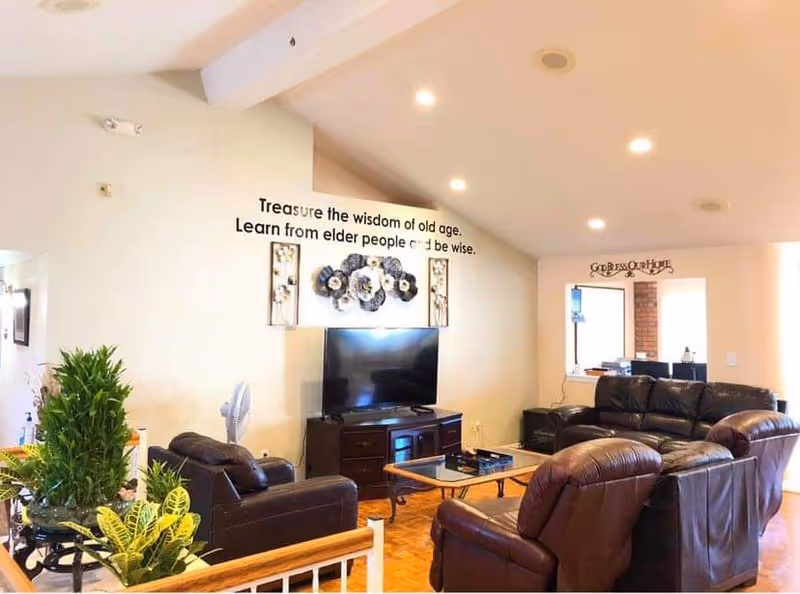 A cozy living room with brown leather sofas and armchairs arranged around a glass coffee table. A flat-screen TV sits on a wooden TV stand against the wall, which is decorated with floral wall art and a quote that reads 'Treasure the wisdom of old age. Learn from elder people to be wise.' There are plants in the foreground and a sign above a doorway in the background that says 'God Bless Our Home.' The room is well-lit with recessed ceiling lights.
