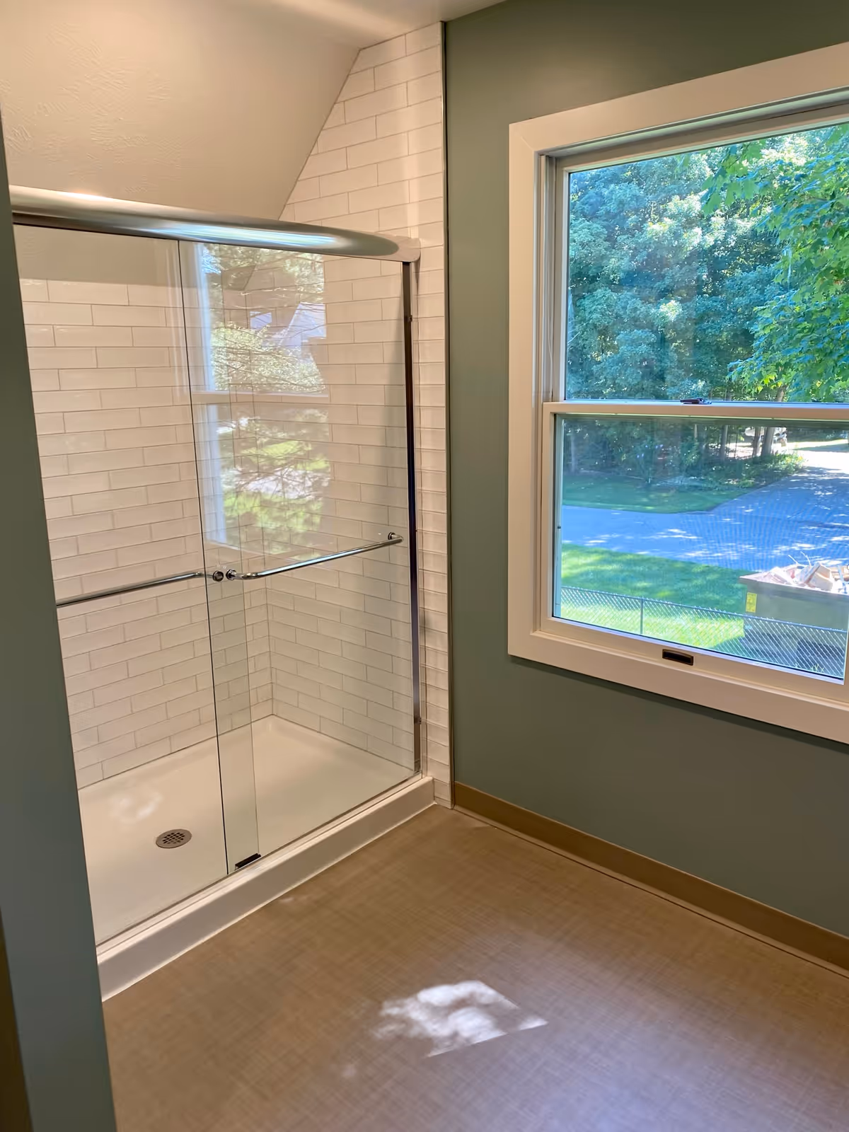 A clean bathroom shower area with white subway tile walls and a glass sliding door. There is a window next to the shower showing green trees and a road outside. The floor is a light brown color and the walls are painted green.