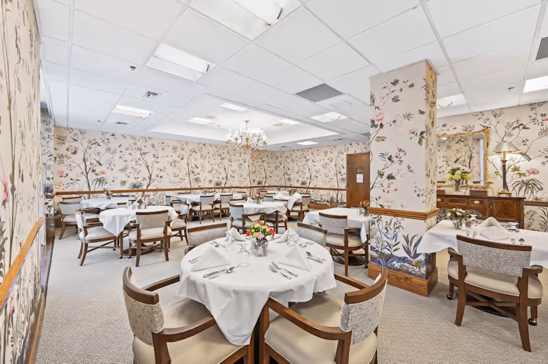 A dining room in Mayfair Village Retirement Community with round and rectangular tables covered in white tablecloths, set with silverware, napkins, and floral centerpieces. The walls are decorated with floral wallpaper, and there is a chandelier hanging from the ceiling. Chairs with wooden frames and cushioned seats surround the tables.