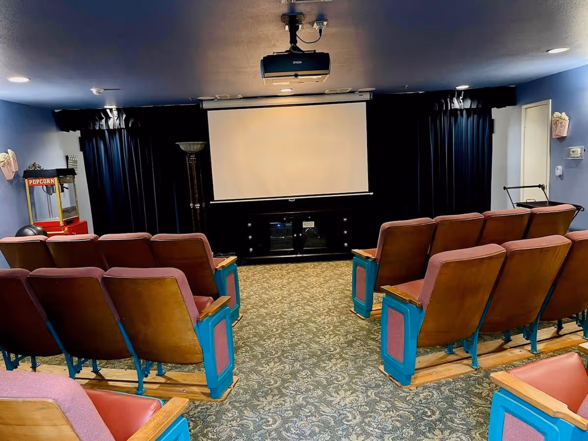 A small theater room with rows of vintage-style seats facing a large white projection screen. The room has patterned carpet, blue walls, and a popcorn machine on the left side. There is a ceiling-mounted Epson projector above the screen and dark curtains behind it.